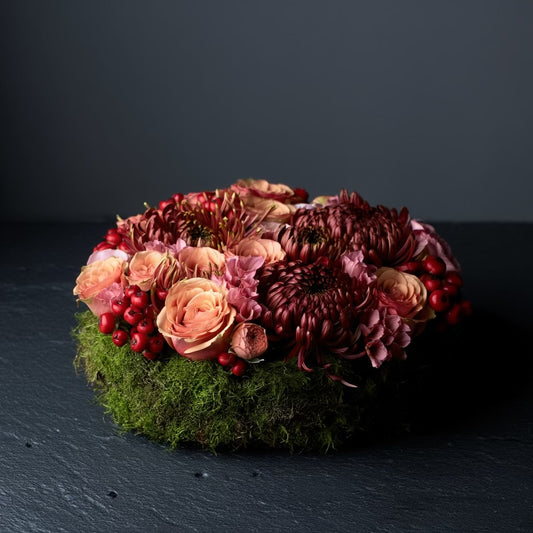 Een rond bloemstuk in taartvorm, afgewerkt met warme, diepe tinten bordeaux en zalm, en omgeven door een mosrand. Chrysanten, rozen, rozenbottels, lycianthus.