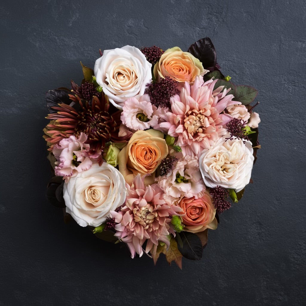 Een rond bloemstuk in taartvorm, afgewerkt met warme, diepe tinten bordeaux en zalm, en omgeven door een mosrand. Chrysanten, rozen en lycianthus.