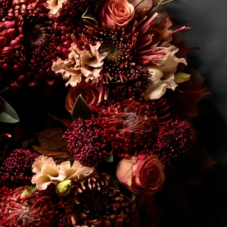 De krans is opgebouwd uit rode Leucospermum (speldenkussenprotea), roestkleurige Chrysanten en Rozen, aangevuld met Skimmia ter opvulling.