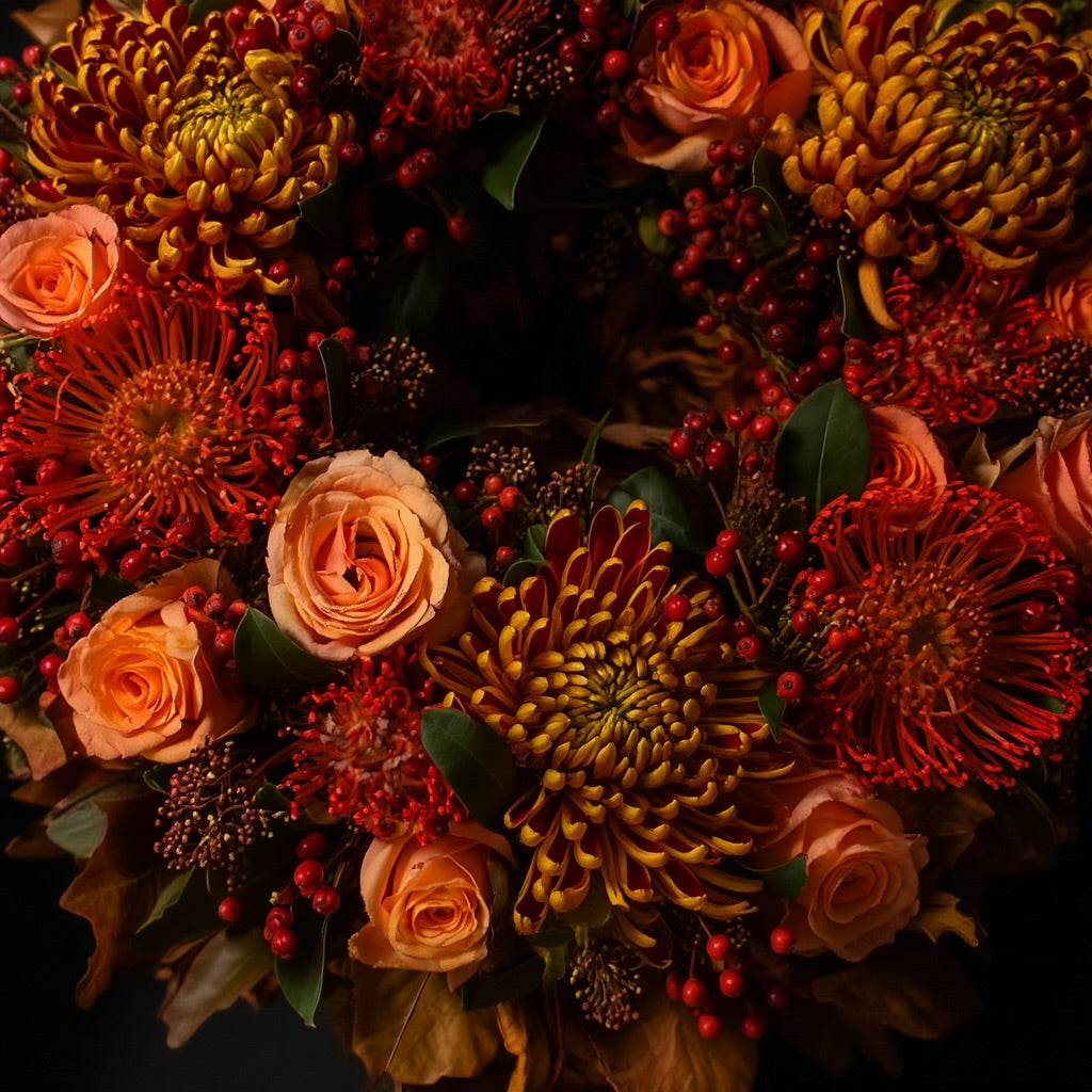 De krans is opgebouwd uit rode Leucospermum (speldenkussenprotea), roestkleurige Chrysanten en oranje-roze Rozen, aangevuld met Skimmia.