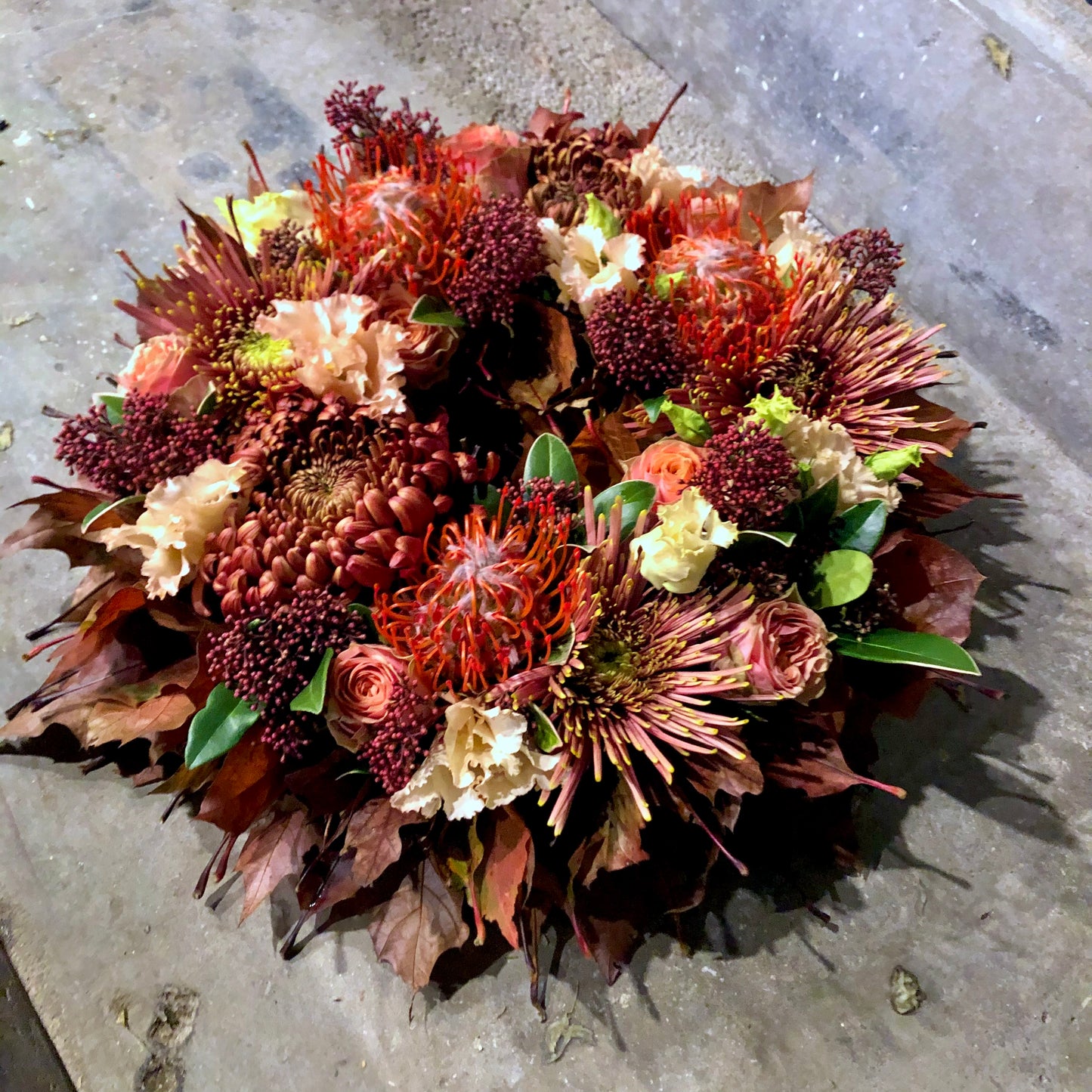 Allerheiligen bloemenkrans in herfsttinten gevuld met rozen, chrysanten, protea’s, bessen afgewerkt met herfstbladeren