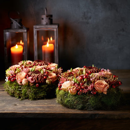 Een rond bloemstuk in taartvorm, afgewerkt met warme, diepe tinten bordeaux en zalm, en omgeven door een mosrand. Chrysanten, rozen, rozenbottels, lycianthus.