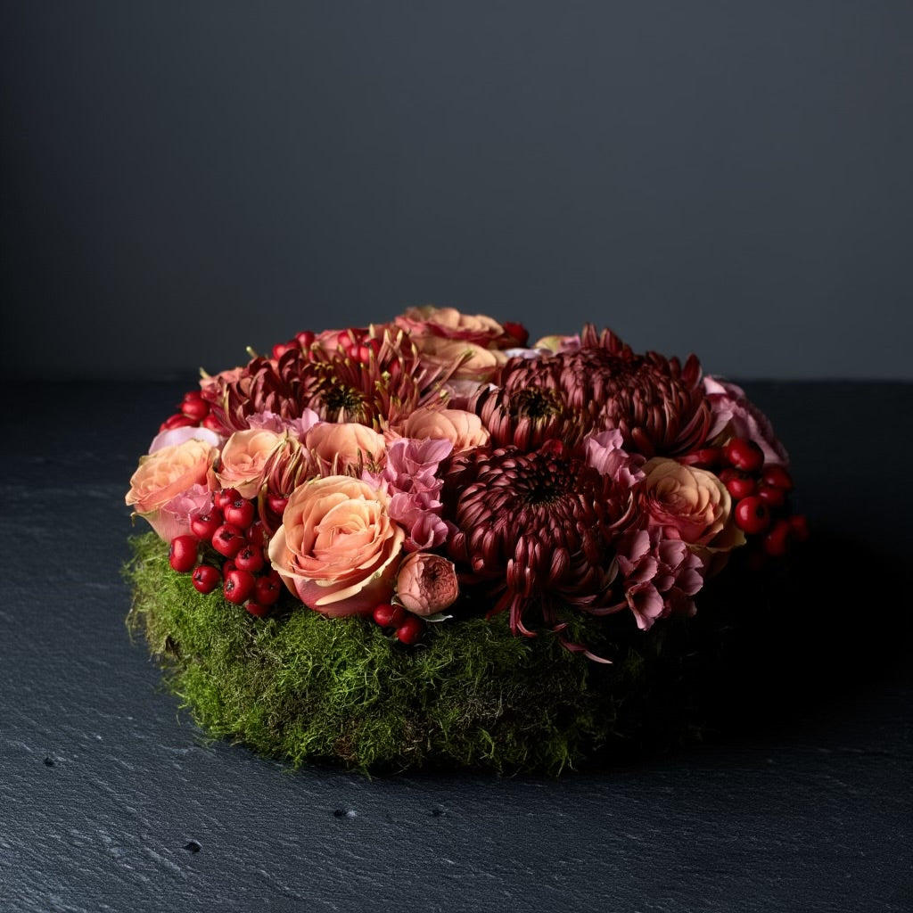 Een rond bloemstuk in taartvorm, afgewerkt met warme, diepe tinten bordeaux en zalm, en omgeven door een mosrand. Chrysanten, rozen, rozenbottels, lycianthus.