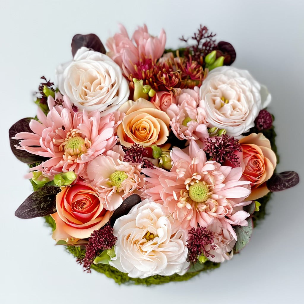 Een rond bloemstuk in taartvorm, afgewerkt met warme, diepe tinten bordeaux en zalm, en omgeven door een mosrand. Chrysanten, rozen en lycianthus.