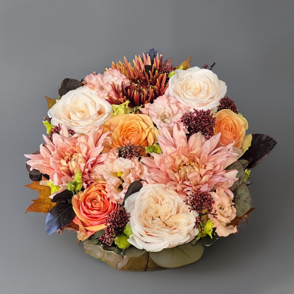 Een rond bloemstuk in taartvorm, afgewerkt met warme, diepe tinten bordeaux en zalm, en omgeven door een mosrand. Chrysanten en soorten rozen.
