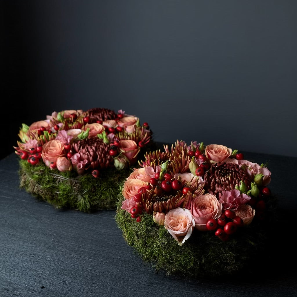 Een rond bloemstuk in taartvorm, afgewerkt met warme, diepe tinten bordeaux en zalm, en omgeven door een mosrand. Chrysanten, rozen, rozenbottels, lycianthus.