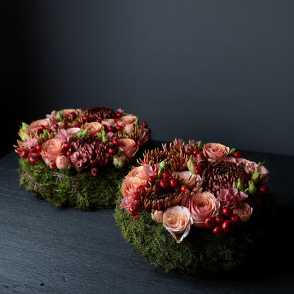 Een rond bloemstuk in taartvorm, afgewerkt met warme, diepe tinten bordeaux en zalm, en omgeven door een mosrand. Chrysanten, rozen, rozenbottels, lycianthus.