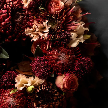 De krans is opgebouwd uit rode Leucospermum (speldenkussenprotea), roestkleurige Chrysanten en Rozen, aangevuld met Skimmia ter opvulling.