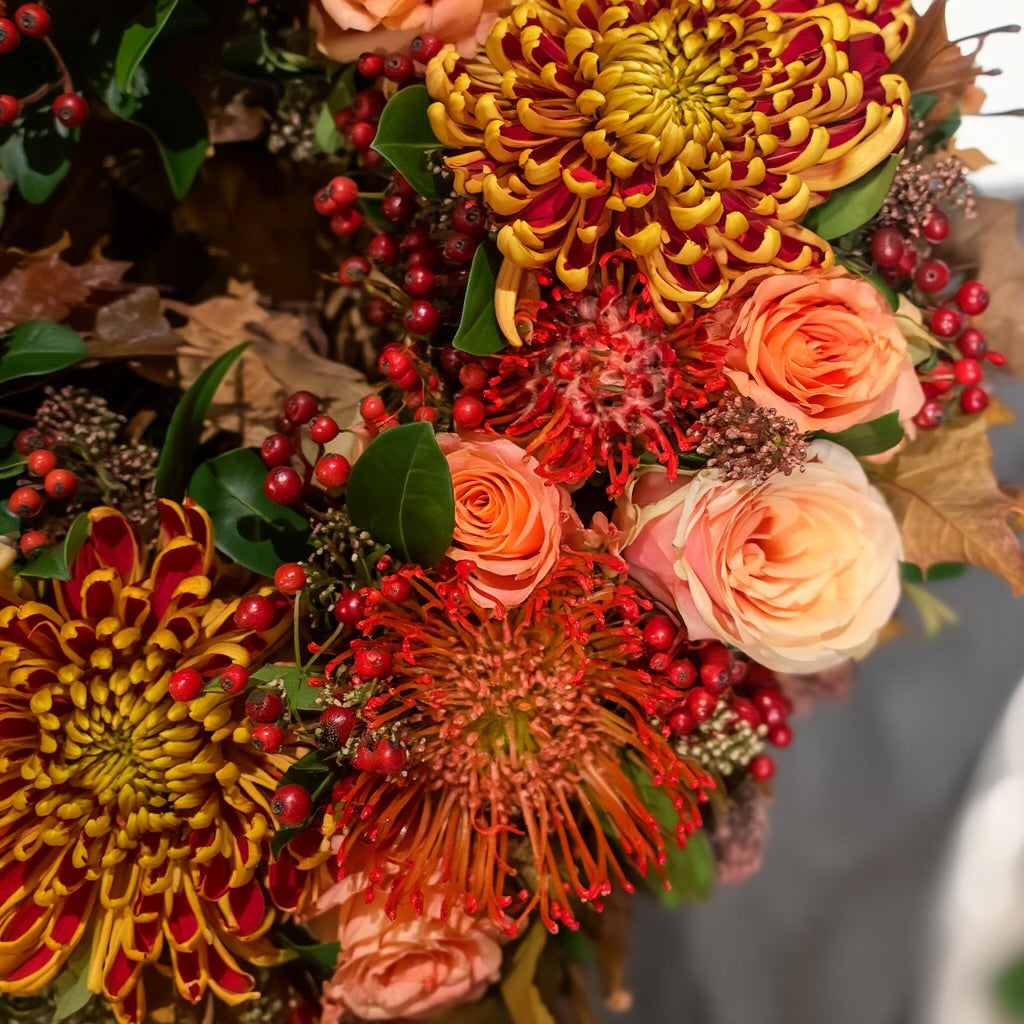 De krans is opgebouwd uit rode Leucospermum (speldenkussenprotea), geel-rode Chrysanten en oranje-roze Rozen, aangevuld met Skimmia.