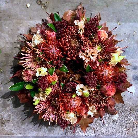 Allerheiligen bloemenkrans in herfsttinten gevuld met rozen, chrysanten, protea’s, bessen afgewerkt met herfstbladeren